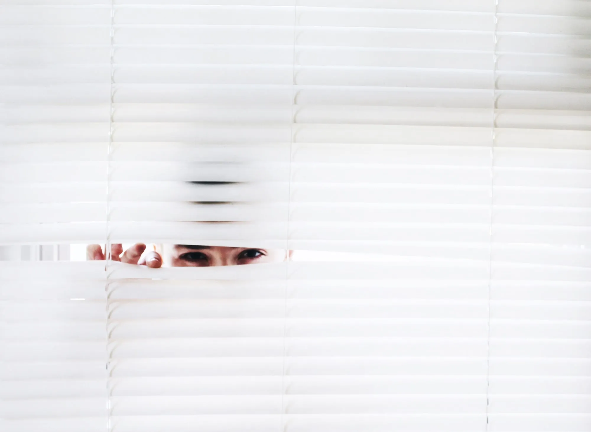 An eye peeking through a gap in white window blinds.