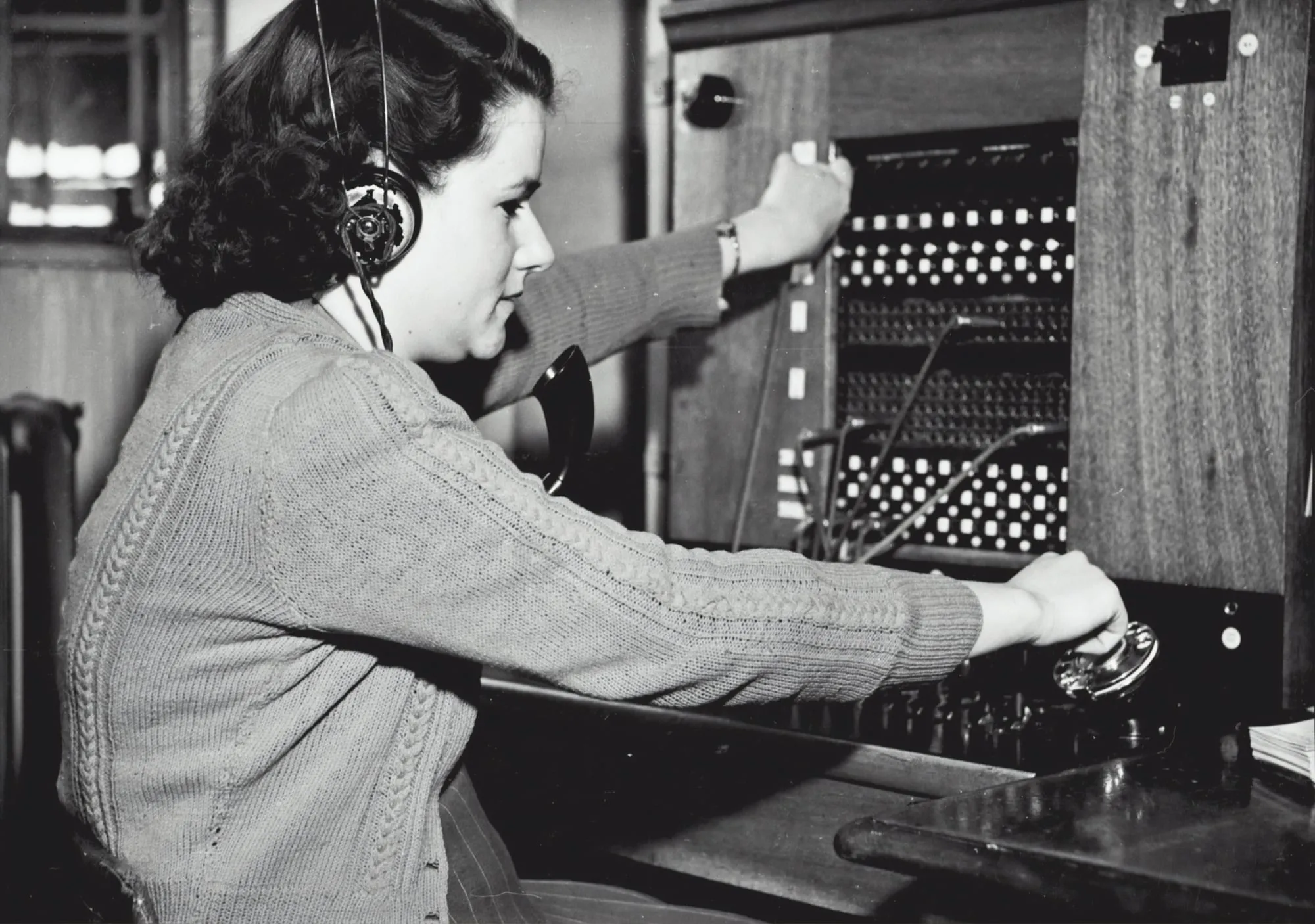 A woman in a headset operating a manual telephone switchboard, in black and white.