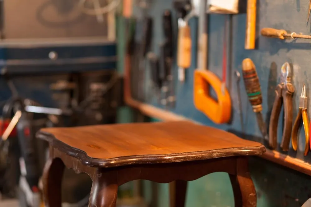 A wooden stool in a workshop with tools hanging on the wall behind it.