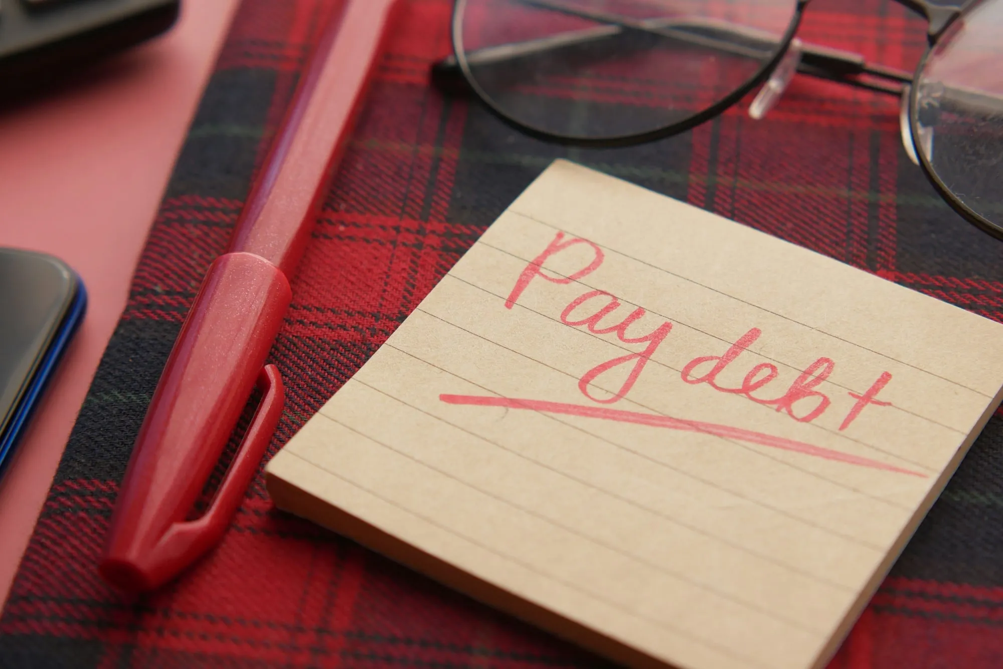 A sticky note reading “Pay debt” beside a red pen and glasses.
