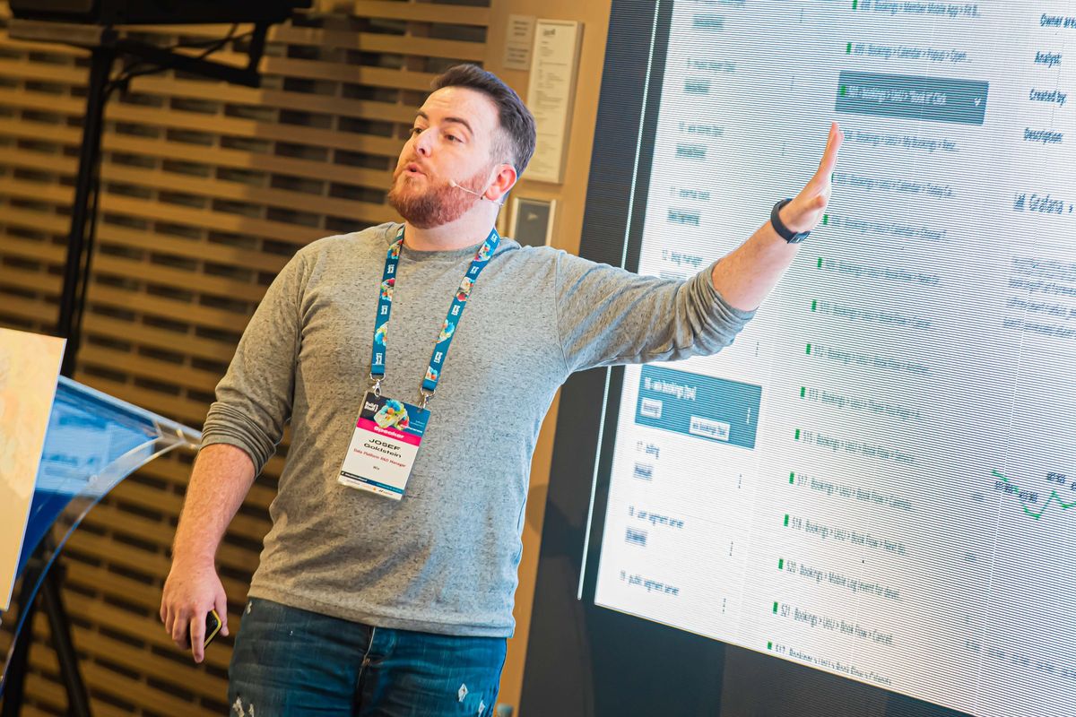 Yossi in a grey sweater and speaker lanyard, arm outstretched toward a screen of event schemas at Build Stuff