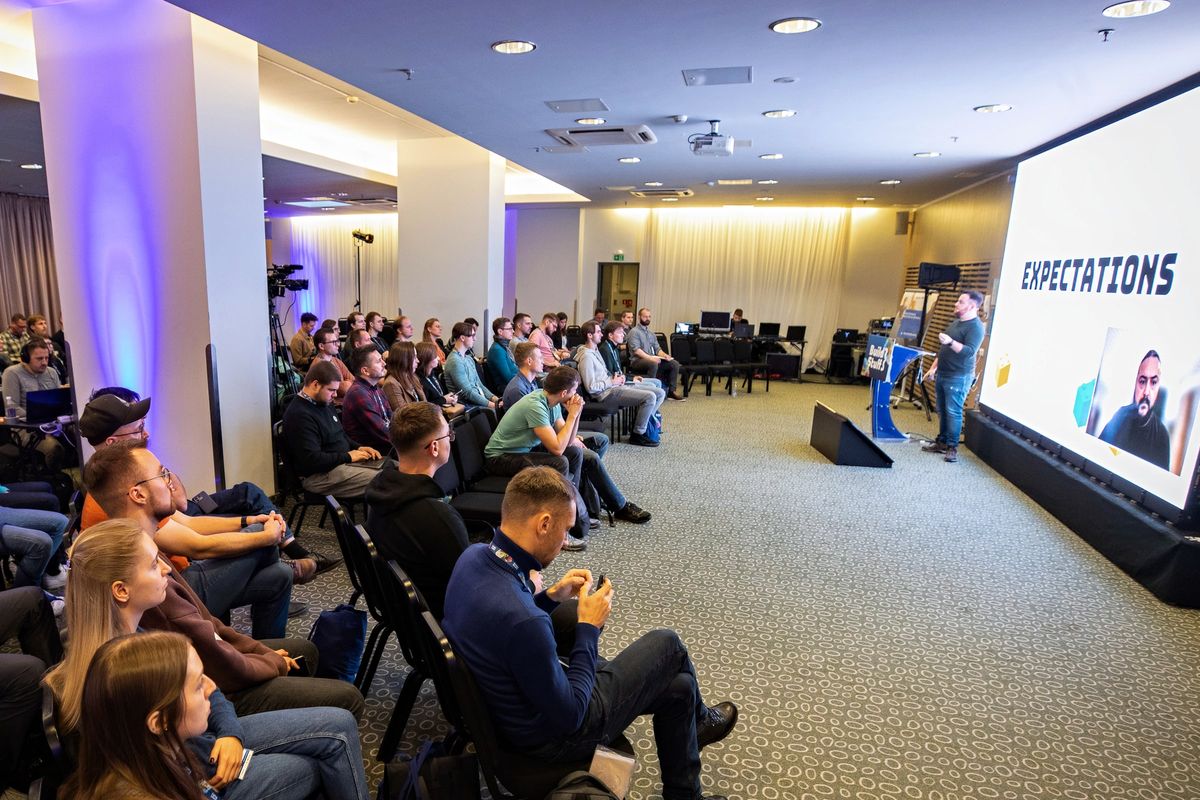 A full room at Build Stuff watching Yossi present beside a giant slide reading EXPECTATIONS, co-speaker David Ostrovsky on the remote feed