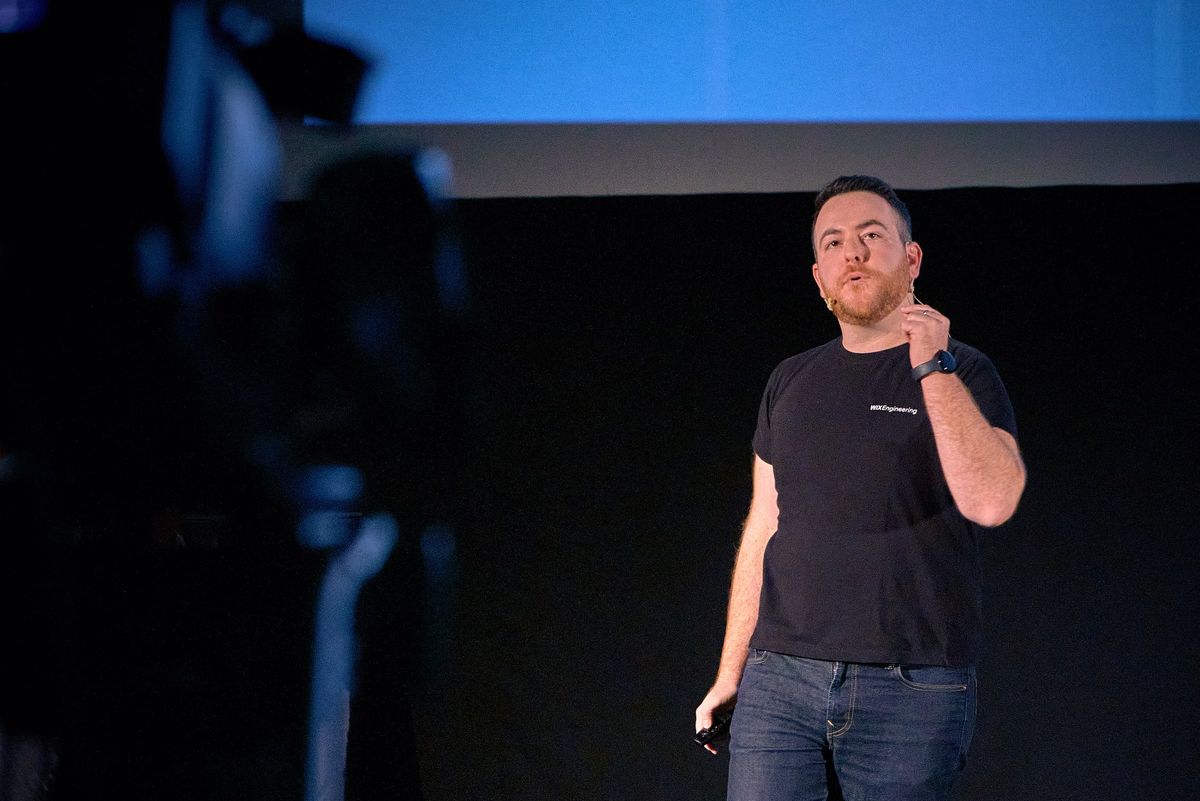 Yossi mid-explanation on a dark stage under a blue screen, a film camera blurred in the foreground