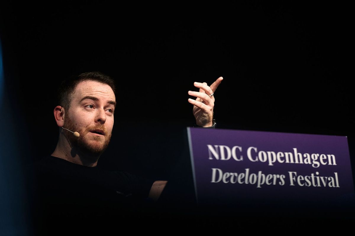 Yossi mid-sentence at the NDC Copenhagen lectern, hand raised, the festival sign glowing purple beside him