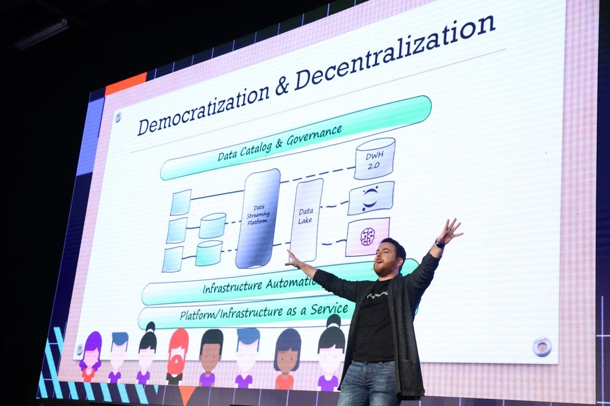 Yossi on the Reversim stage, arms flung wide in front of a slide titled Democratization & Decentralization